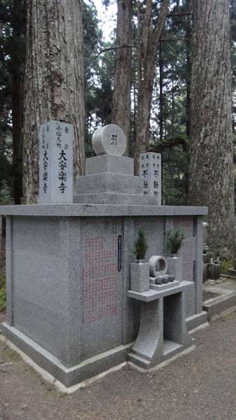 a square gravestone with text marker stones, stacked stone blocks and a circle with kanji on top.  the kanji is reproduced in stone in front of the base.